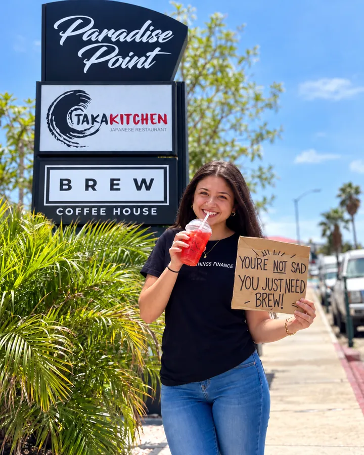 A guest holding a Brew drink and a sign by the Paradise Point sign featuring Brew Coffee House